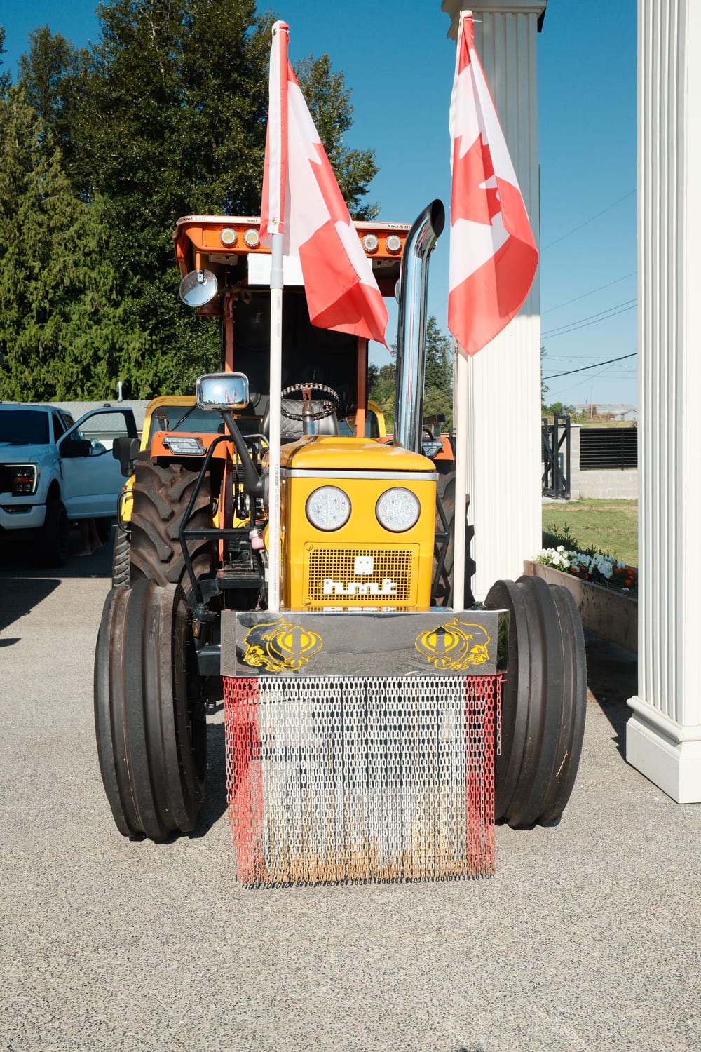 An orange and yellow HMT 5911 show tractor with a chrome exhaust stack, a lamp bar across the roof, and a chain fringe hanging from the front weight bar.